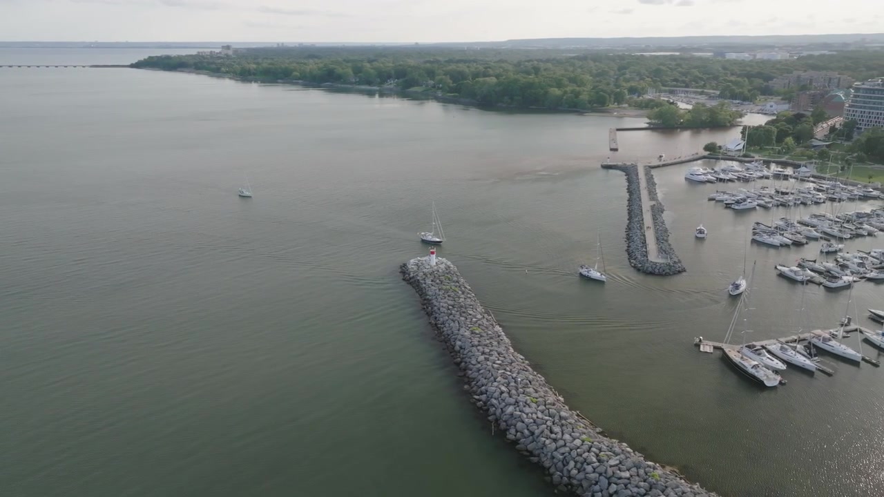 The stone pier stretches toward open water along the Oakville shoreline on Lake Ontario. Parked boats fill the calm marina docks beneath a high hazy summer sky.