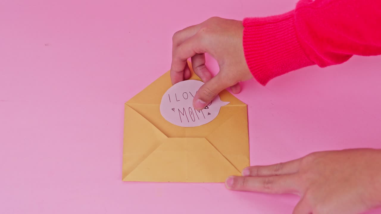 Hand reveals hand-drawn "I Love Mom" note from a classic yellow envelope on pink background