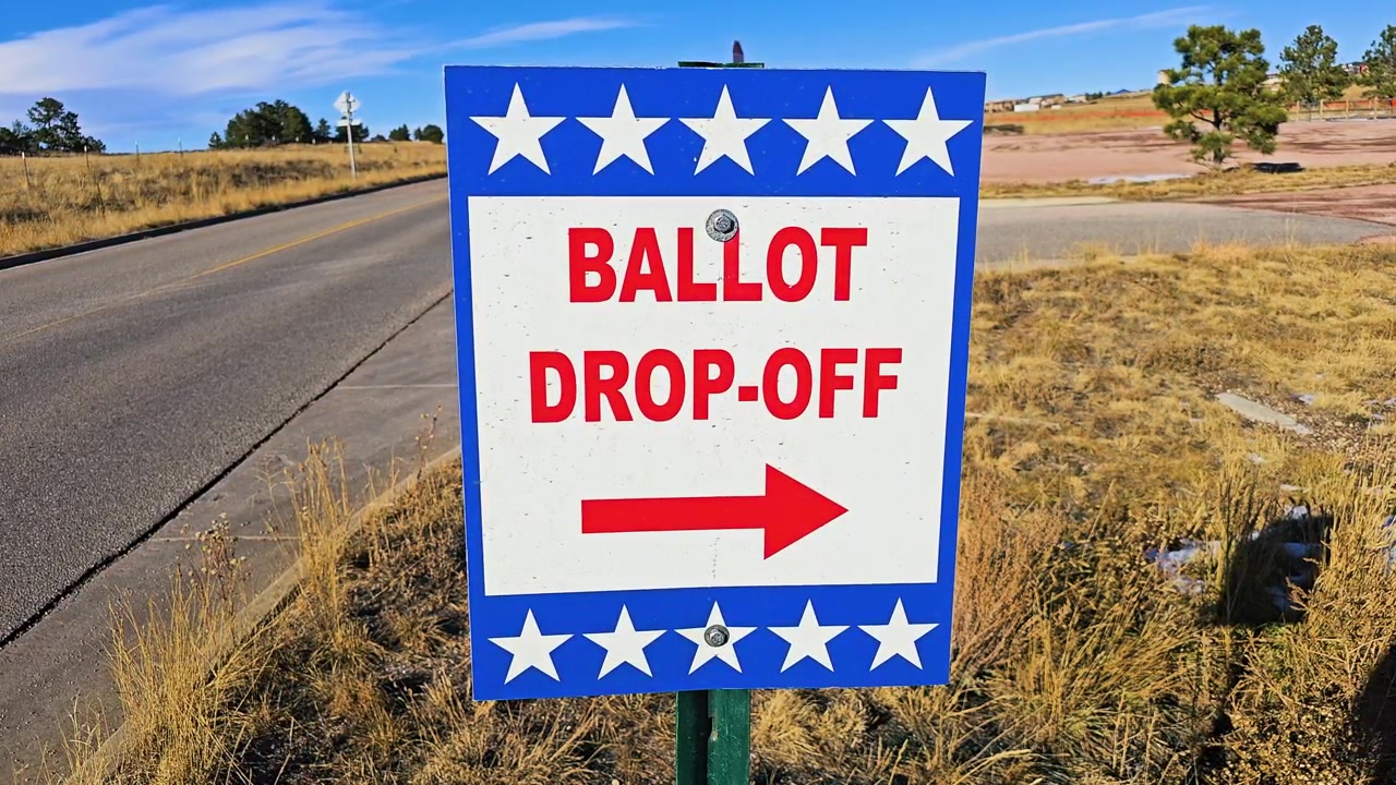 Blue ballot drop-off sign beside a quiet road directs voters with a red arrow, patriotic stars, dry grass, pine trees, and clear blue sky during an American election season outdoors.