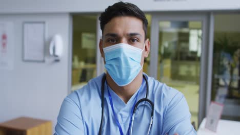Portrait-of-mixed-race-male-doctor-wearing-scrubs-and-face-mask-standing-in-hospital