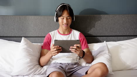 sitting on bed, young asian man using tablet and wearing headphones, smiling