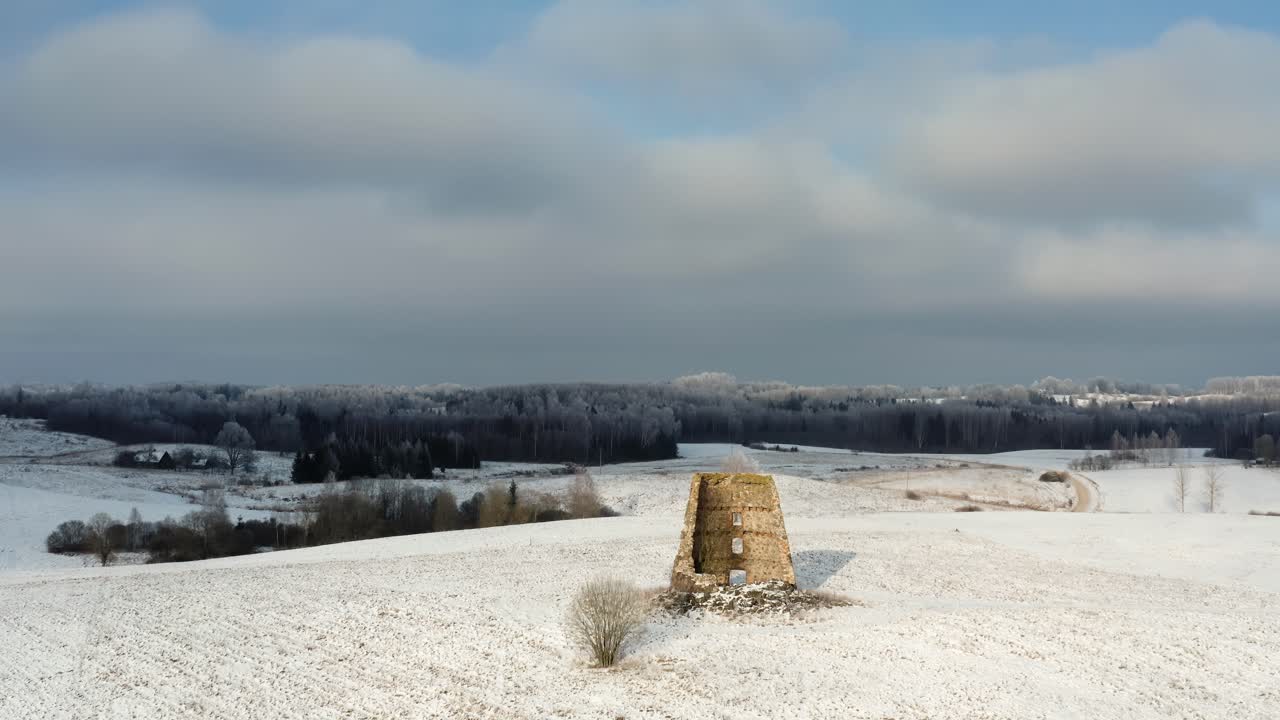 Abandoned windmill ruins on a small snow-covered hilltop. Winter drone shot of frozen forest and sun over the landmark.