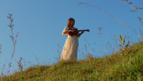 female young asiatic musician artist playing guitar ukulele in nature wearing white long dress