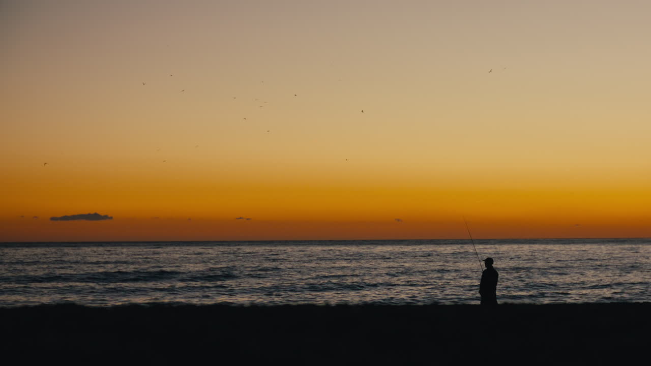 A silhouette of a lone fisherman stands on the beach during sunset, casting his line into the calm sea while a flock of seabirds circles above the golden horizon
