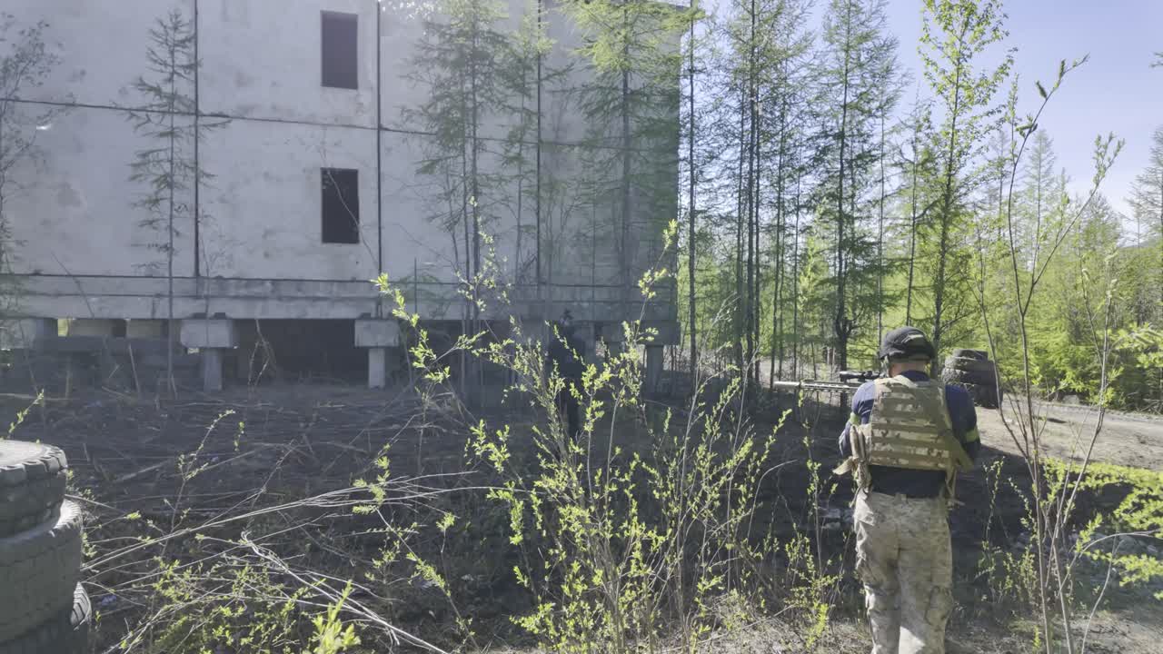 An airsoft player carefully approaches an abandoned building in a wooded area.