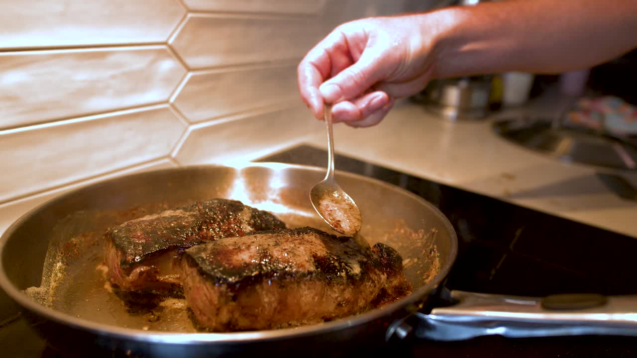 A chef bastes searing beef steaks with melted butter in a stainless steel frying pan