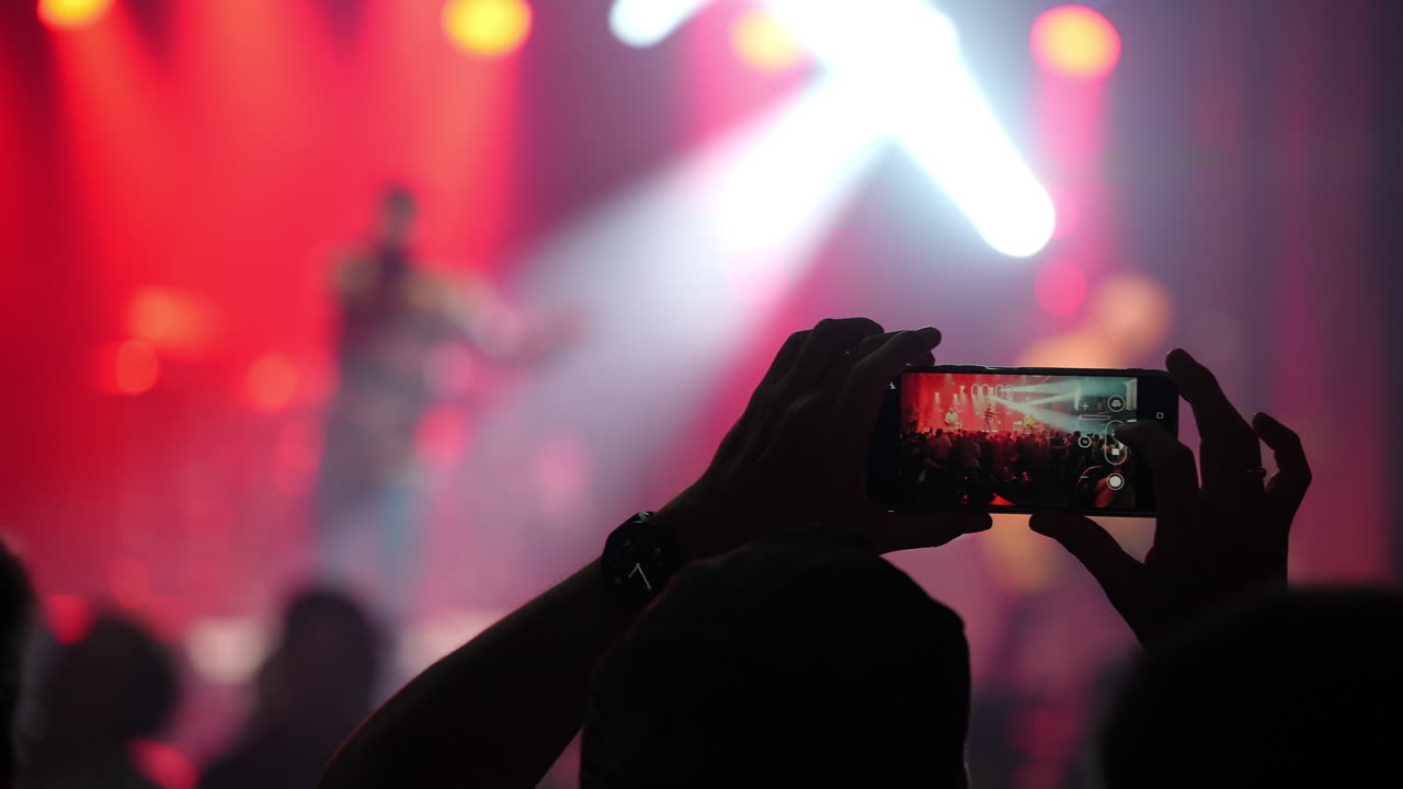 Spettatore che cattura un'esibizione dal vivo con lo smartphone, illuminato da luci rosse vibranti del palco, creando un'atmosfera energica a un evento di concerto musicale