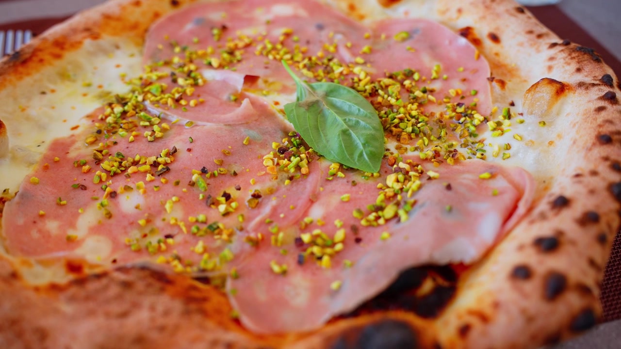 Pizza napoletana cotta a legna di Pompei con mortadella, pistacchi tritati, basilico, formaggio cremoso e un cornicione alveolato su un tavolo di ristorante.