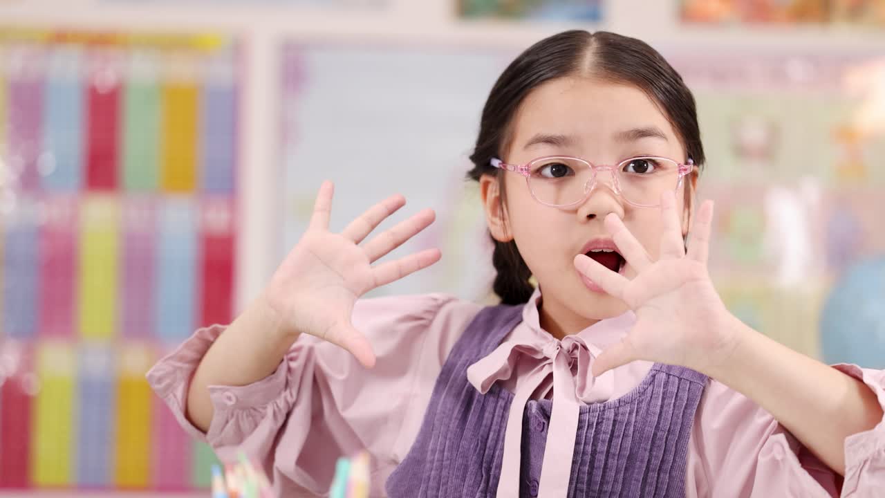Young girl with glasses performs expressive hand gestures in bright, colorful classroom, front-facing camera