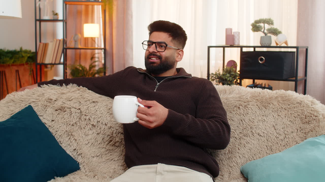 Indian man enters room on sofa enjoys coffee with a smile after stressful day at home with comfort