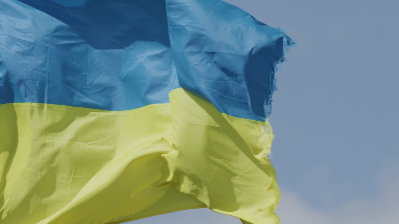 Blue and yellow Ukrainian flag flaps vigorously against a clear sky, captured in close-up with natural daylight and dynamic camera movement