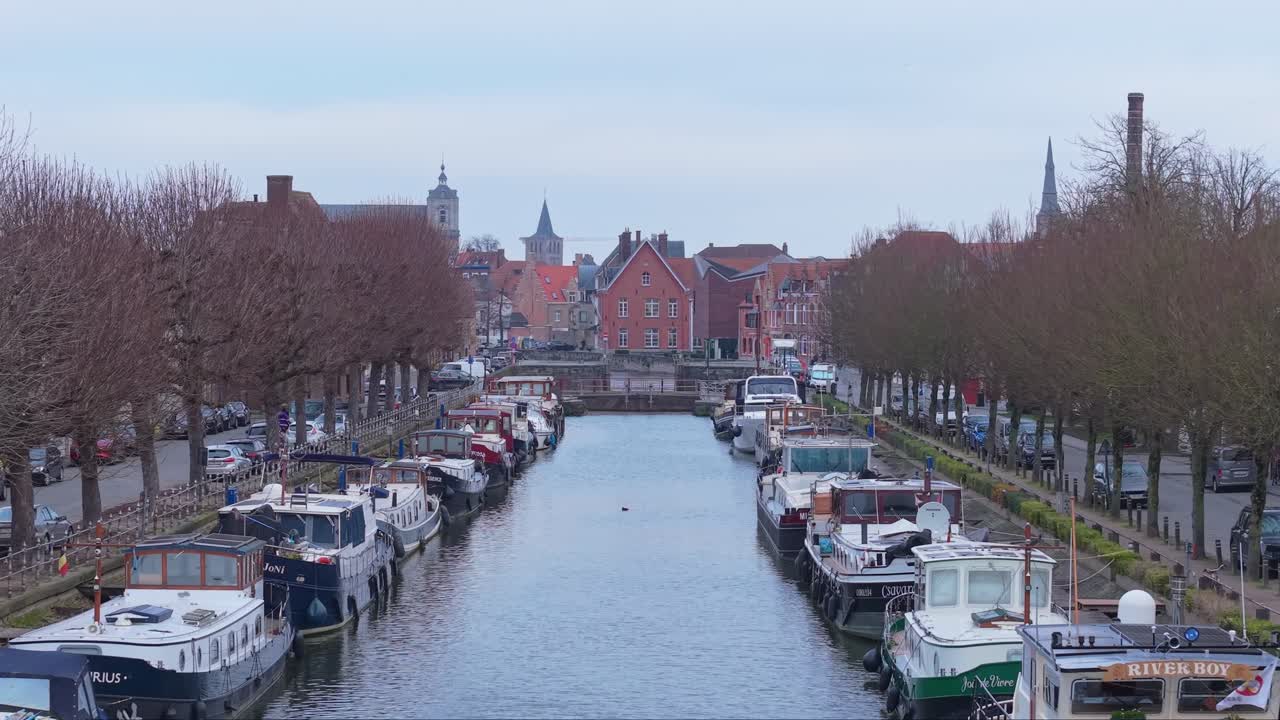 Una ripresa aerea cinematografica sorvola il canale di Bruges, rivelando la riva del fiume, i parchi verdi, le imbarcazioni fluviali e le case affascinanti lungo il litorale, catturando il pittoresco paesaggio urbano medievale