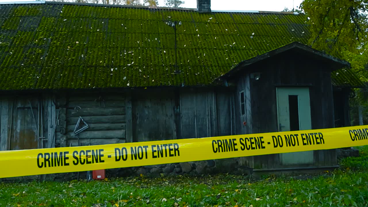 Police yellow colored crime scene forensic investigation tape ribbon line pulled in front of a rural countryside farm house or shed with a eternit roof and green moss on it. Autumn cloudy day time