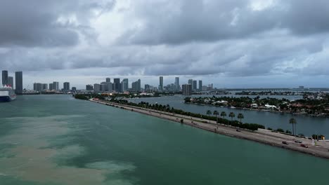 a busy highway surrounded by water at the popular cruise port in miami, florida with dirt in the water from the ship moving and a cruise ship and skyscrapers in the background on a warm overcast day