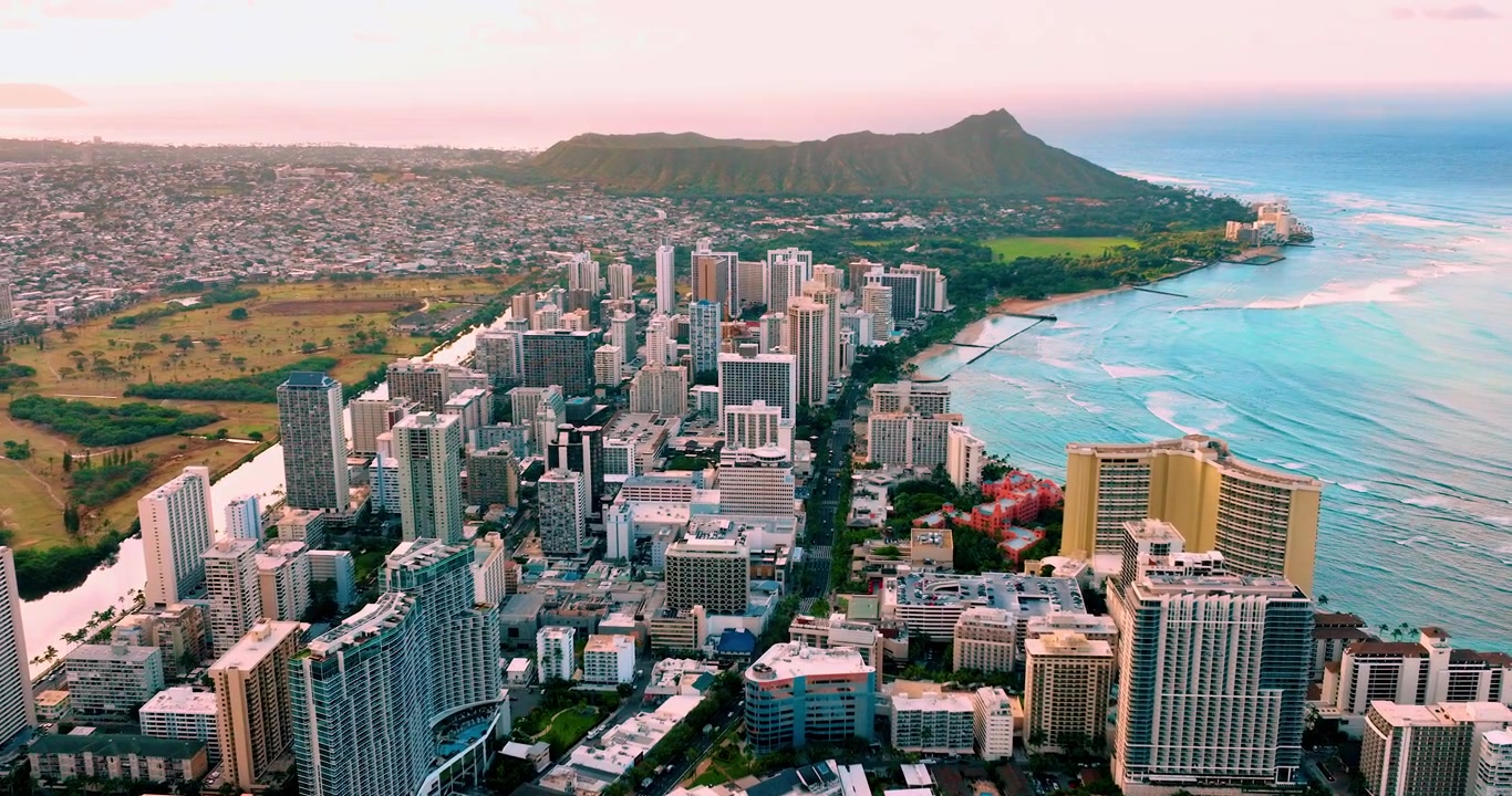 Bagliore del tramonto sul quartiere degli hotel di Waikiki e sul cratere Diamond Head. Vista aerea di un denso gruppo di torri resort accanto alla costa turchese, sagoma del cratere vulcanico a destra, cielo rosa e blu al crepuscolo