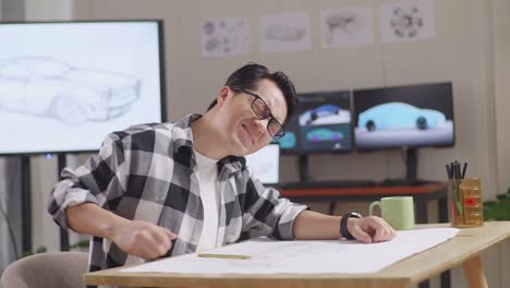 side view of asian male having a backache while working on a car design sketch on table in the studio with tv and computers display 3d electric car model