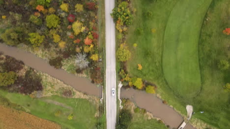 aerial top down tracking view of white car vehicle driving moving along rural countryside road, route through nature autumn fall season landscape, green lands fields and colorful trees around