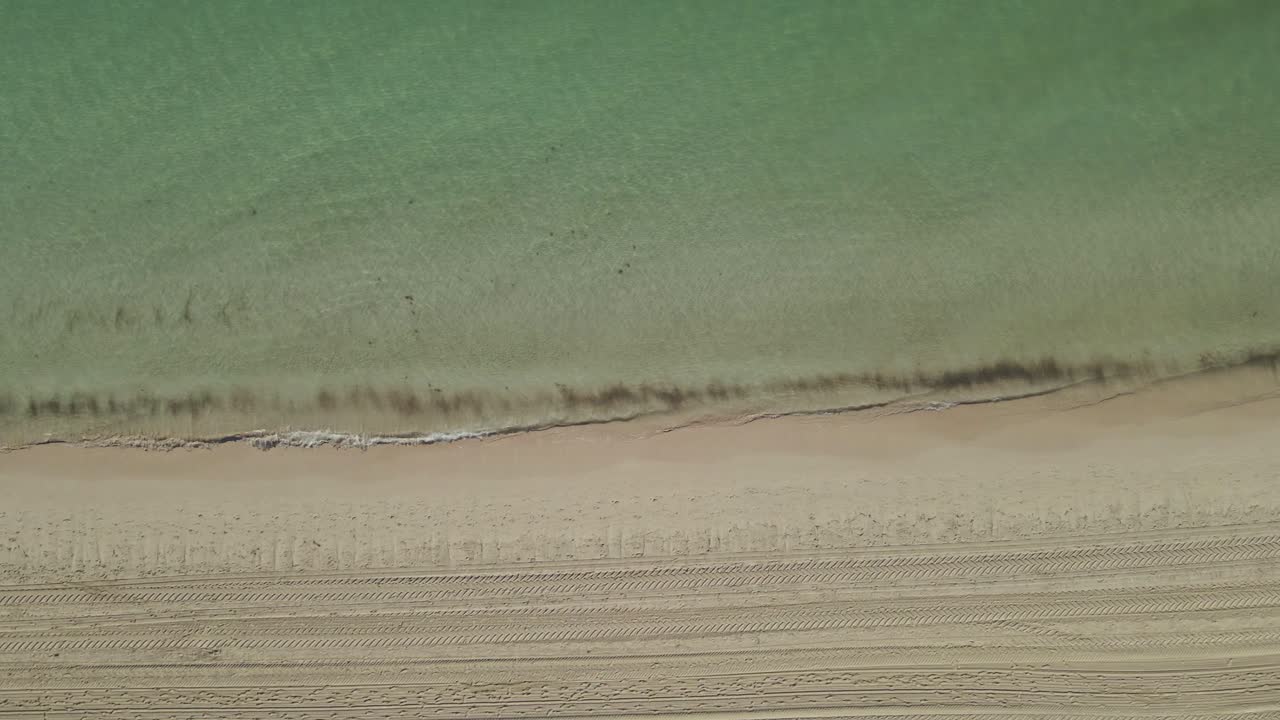 A top-down aerial view of a pristine tropical beach with clear turquoise water gently washing onto smooth, golden sand.