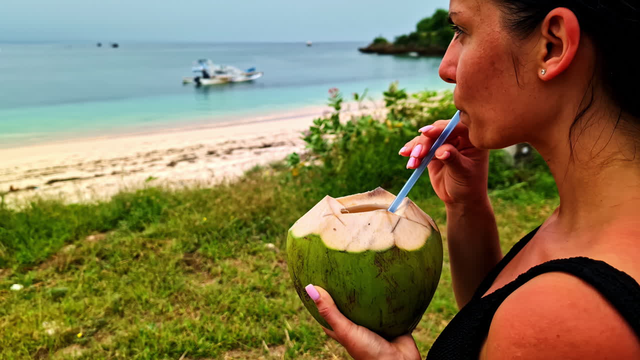 A beautiful young woman drinks from a fresh green coconut with a straw, enjoying the view of the famous Pink Beach on a relaxing tropical holiday in Lombok, Indonesia