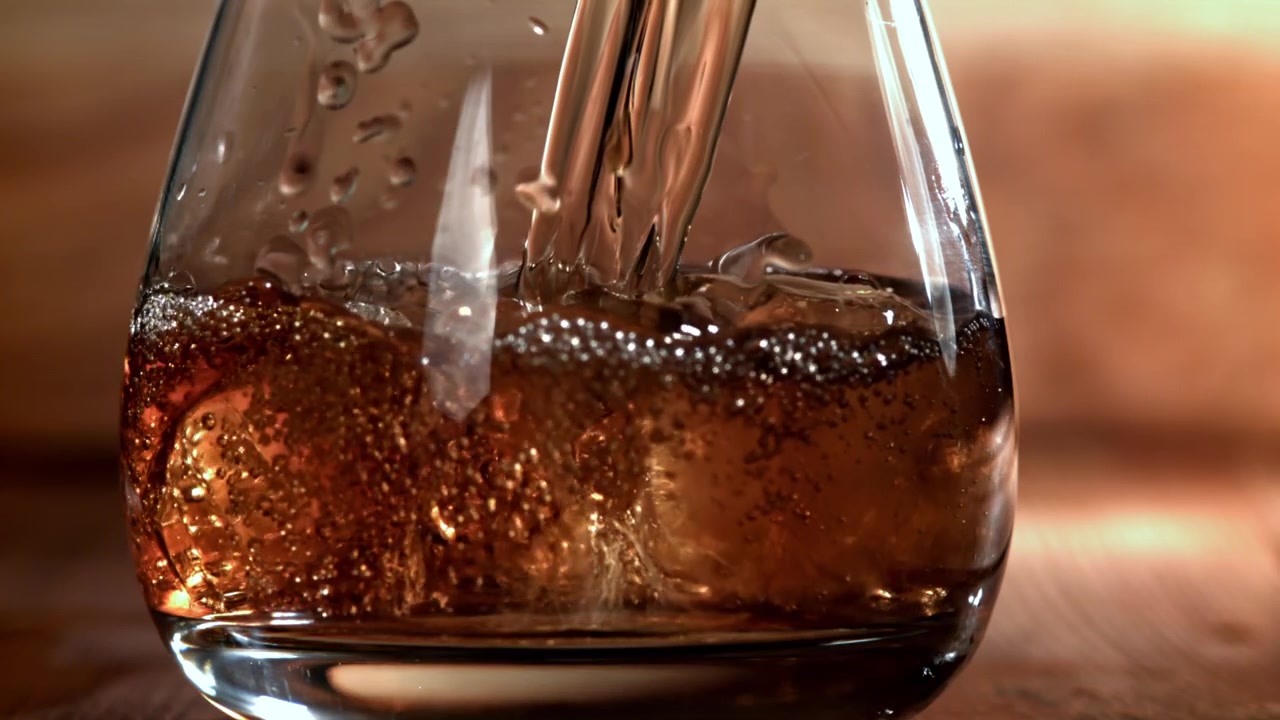Versamento al rallentatore di una bevanda ambrata rinfrescante con ghiaccio in un bicchiere