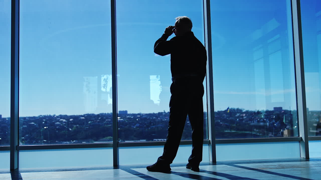Silhouette of a senior man wearing glasses standing at the big panoramic window in office. Man takes off his glasses and puts them again. Low angle view.