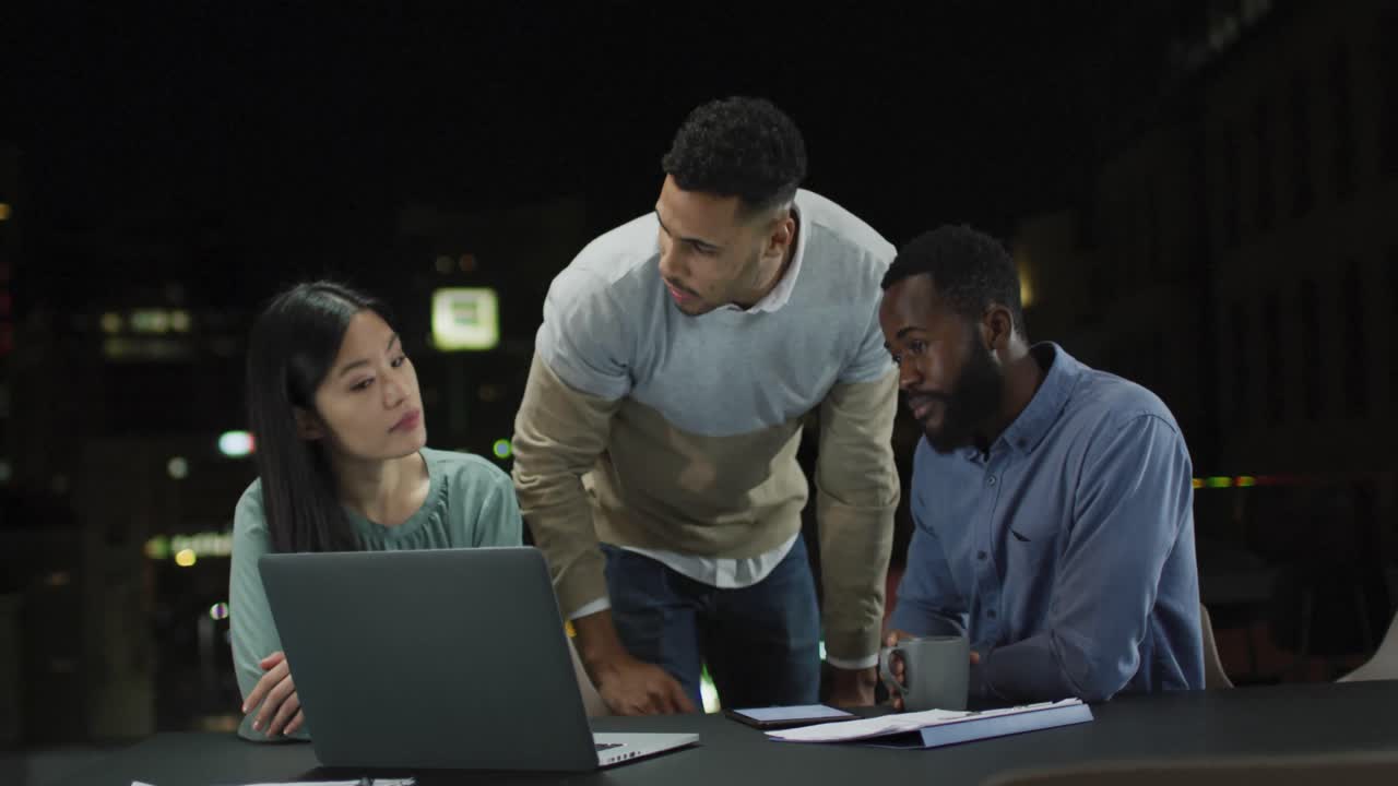 Video of three diverse male and female colleagues in discussion around a laptop at night in office