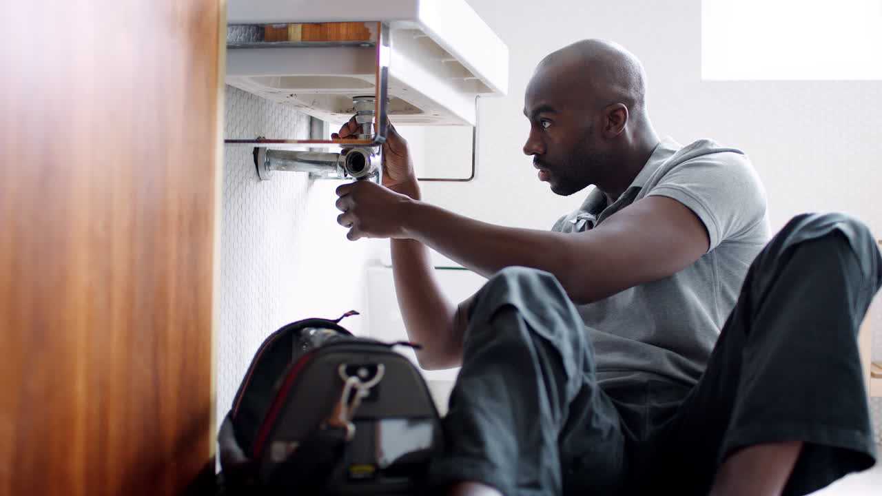 Young black male plumber sitting on the floor and replacing the trap pipe under a bathroom sink, seen from doorway