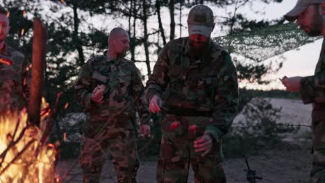 the soldiers are resting after their service in field they are warming themselves at hearth with bonfire, they are cutting sausages frying them on sticks, preparing supper, darkness falls after sunset