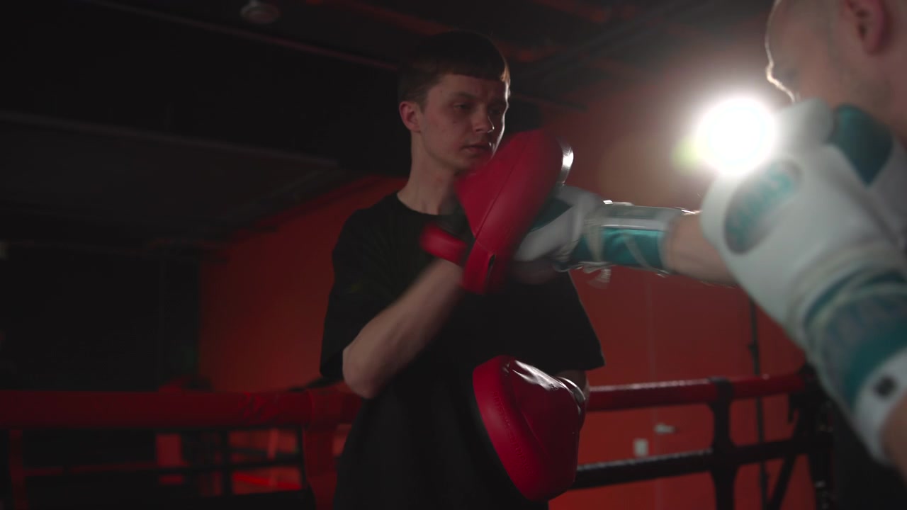Un pugile in primo piano e il suo partner si esercitano in allenamenti mirati con i colpitori in un ring da palestra buio, con colpitori rossi, il bagliore luminoso di una finestra, pareti industriali, sacchi da boxe appesi e un movimento atletico concentrato.