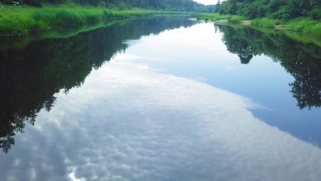 aerial view of a venta river on a sunny summer day, lush green trees and meadows, beautiful rural landscape, wide angle drone shot moving backward low over the calm river