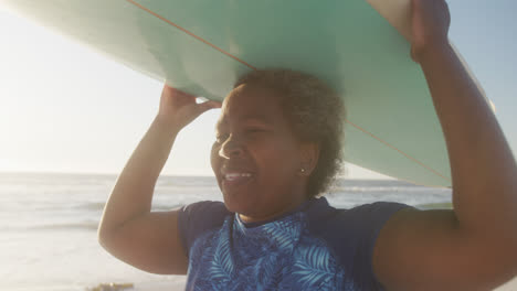 happy senior african american woman walking and holding surfboard at beach, in slow motion