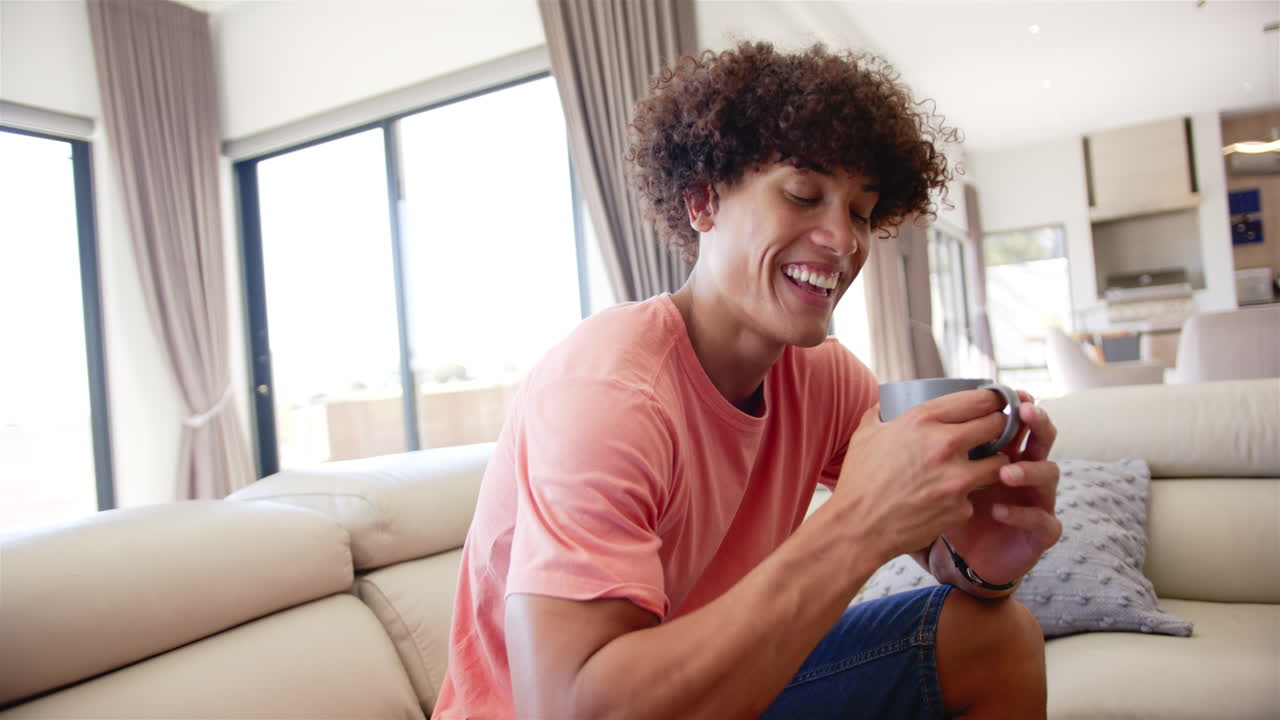 A young biracial man is smiling at his smartphone on the couch at home