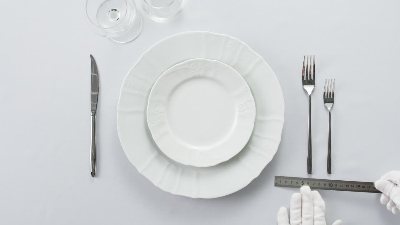 Professional table setting with white gloves. Plates, fork, knife, and glass arranged on a white table.