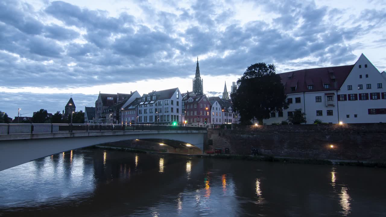 A captivating timelapse of Ulm cityscape during sunset, featuring a bridge in the foreground and the warm hues of the setting sun illuminating the urban skyline.