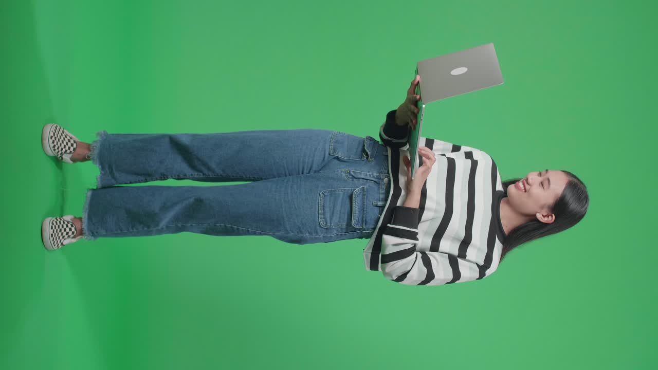 cuerpo lleno de una mujer asiática sonriente escribiendo en la computadora mientras está de pie frente al fondo de la pantalla verde