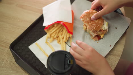 hand view of a woman dropping a partially eaten burger with her right hand on a napkin in a tray, the tray contains fries and a coffee cup on a wooden table, she takes a fry and takes a bite