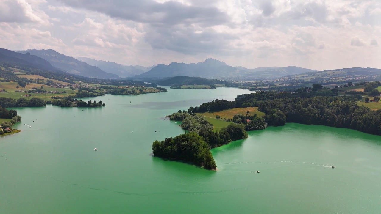 Eine Luftaufnahme einer Drohne zeigt den Greyerzersee bei Pont-la-Ville, Schweiz, mit kleinen bewaldeten Inseln, türkisfarbenem Wasser, entfernten Booten, Ackerland und Bergkämmen unter aufgelockerten Wolken.