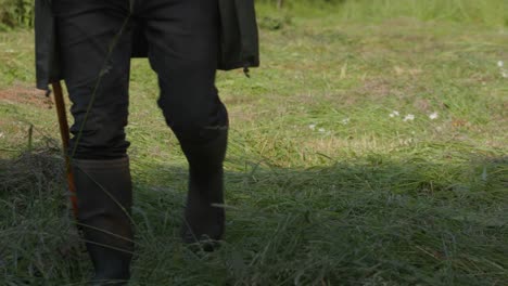 person in rubber boots with wooden stick walks on grass in the field