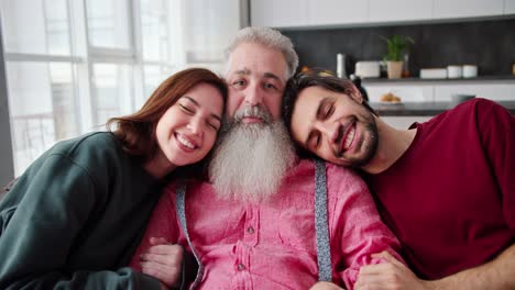 retrato de un hombre feliz con cabello gris y una barba exuberante en una camisa rosa junto con sus hijos adultos un hombre moreno y una chica en una chaqueta verde en un apartamento moderno
