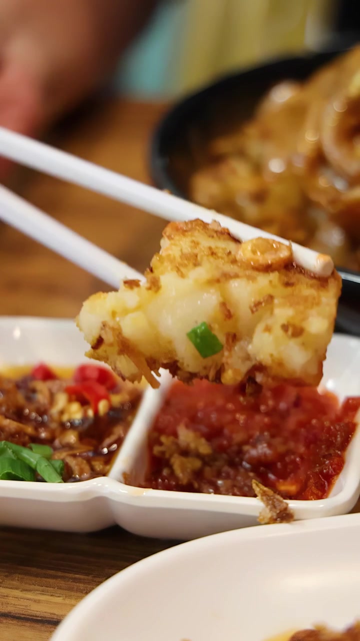 Pemandangan jarak dekat sumpit mengambil sepotong kue lobak goreng, mencelupkannya ke aneka saus di atas meja kayu di bawah pencahayaan dalam ruangan yang hangat.