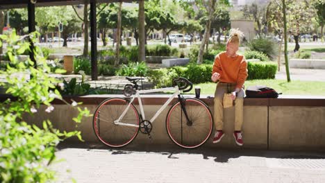 Thoughtful-albino-african-american-man-with-dreadlocks-sitting-in-park-with-bike-using-smartphone