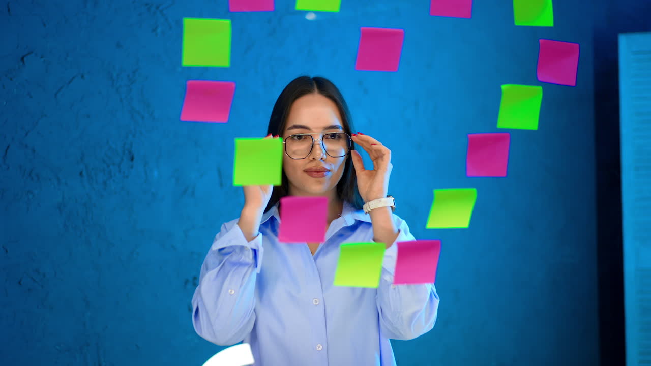 Brunette girl in white shirt and glasses stands at the glass wall. Positive smiling lady touches the sticker notes on the board put in a shape of heart.