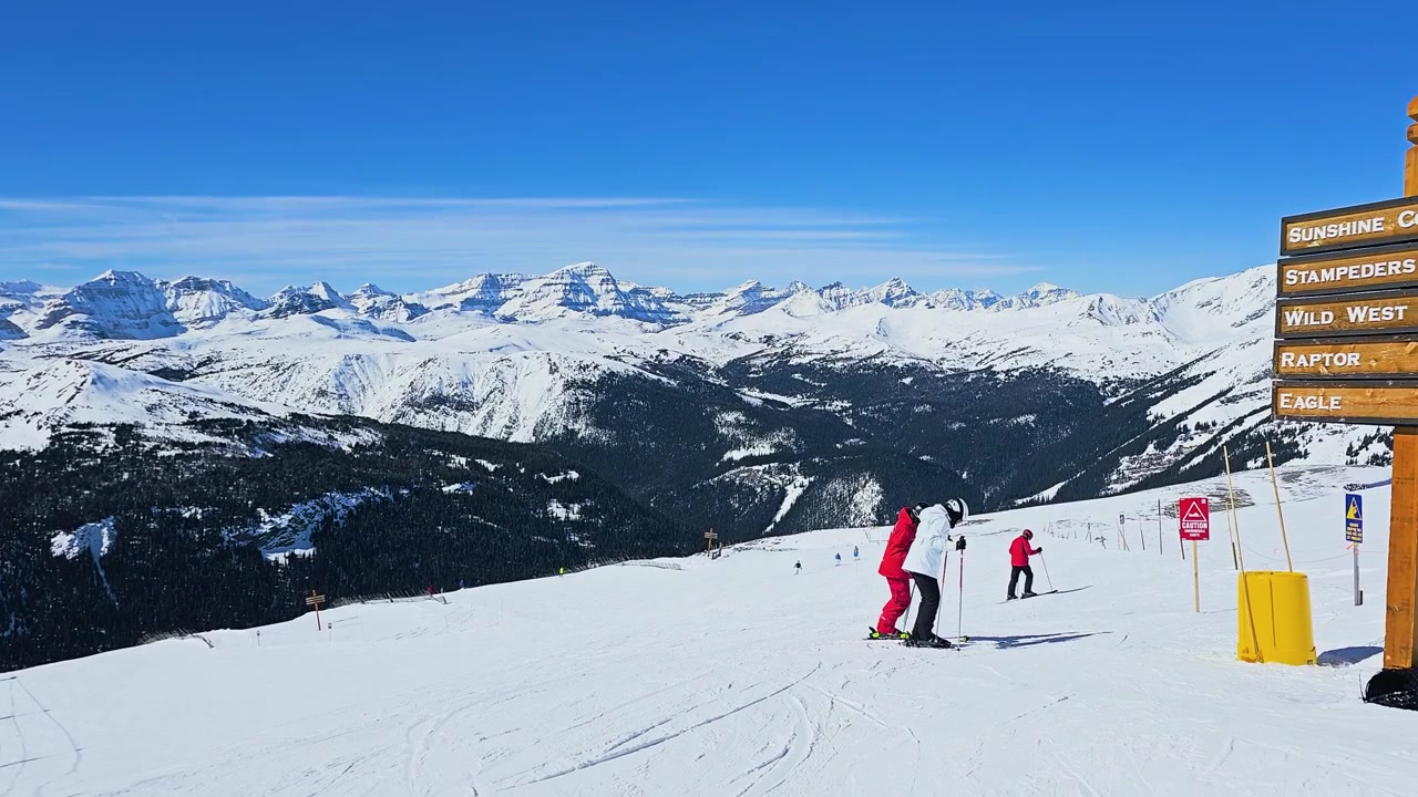 People ski down a groomed mountain run beside a chairlift, capturing an active winter resort scene with panoramic snowy peaks, crisp alpine conditions, and clear blue sky overhead.