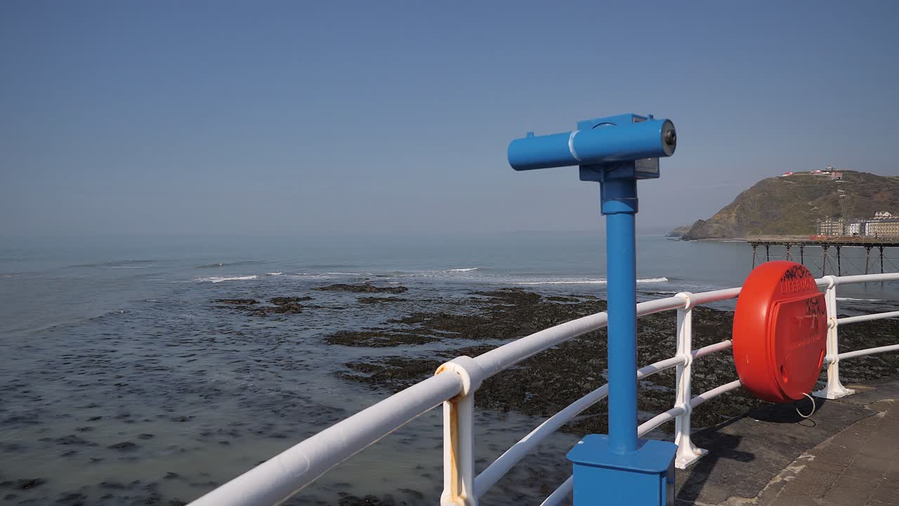 A sea viewing telescope points out to the Irish Sea on the Promenade, Aberystwyth, Ceredigion, Wales on a beautiful clear day. Also on view in the shot is a plastic cover for a life saver ring.
