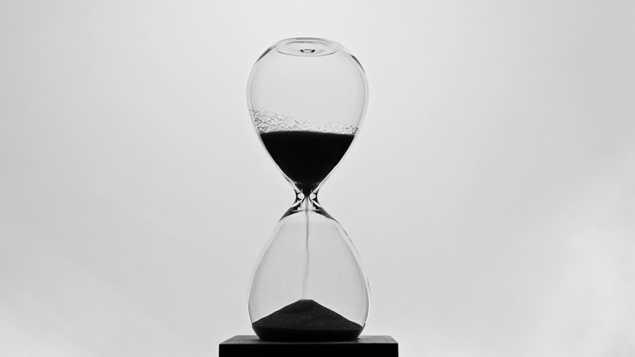 A minimalist black-and-white video of an hourglass with black sand flowing through its narrow neck. The transparent glass and clean white background symbolizing the passage of time.