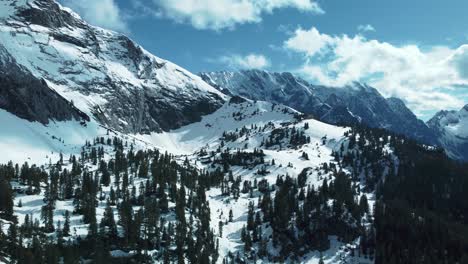 Wide-epic-mountain-panorama-flight-high-above-scenic-Schachen-near-Bavaria-Elmau-castle-and-snowy-glacier-peaks-in-the-alps-on-a-cloudy-and-sunny-day-along-trees,-rocks,-forest-and-hills-in-nature