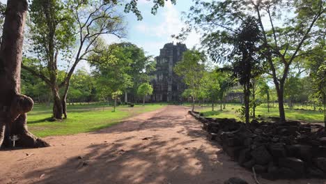 walkthrough shot moves along a path, framed by dense jungle, before unveiling the towering stone pyramid of prasat tom at koh ker, cambodia.