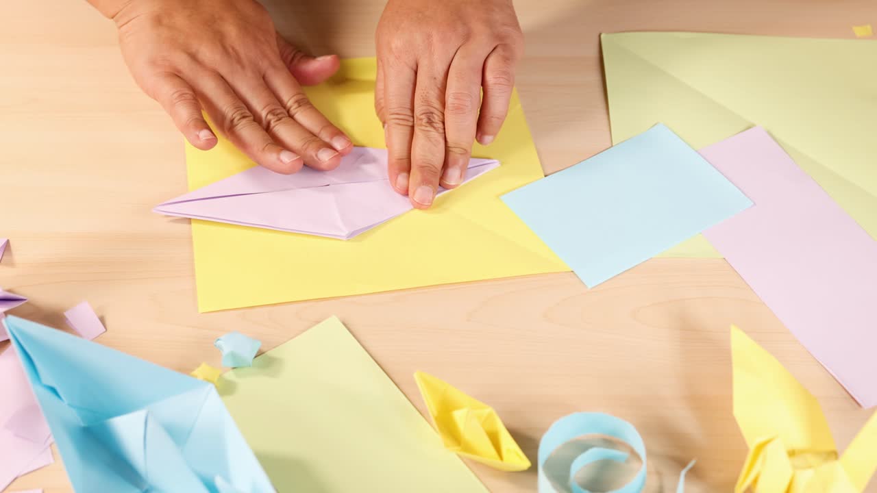Adult hands fold pastel origami boats on wooden table, surrounded by colorful paper, top-down view