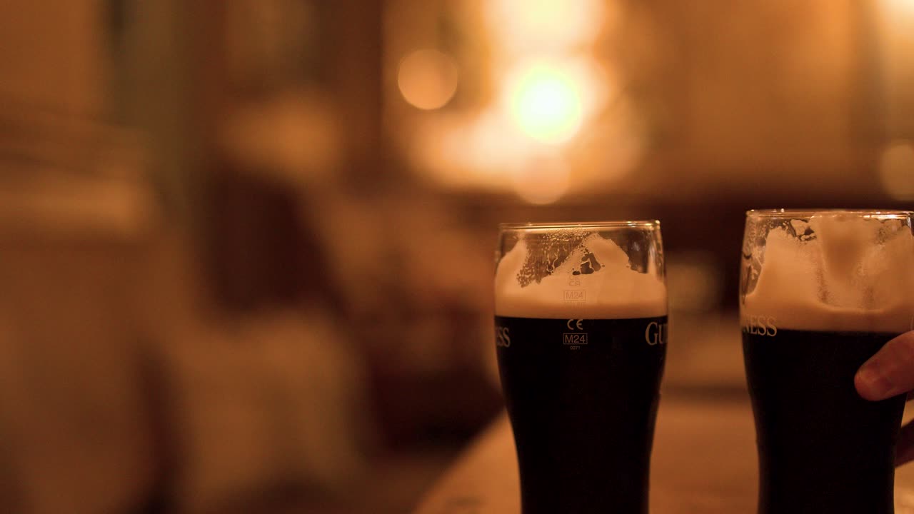 Hand grabs pint of dark beer on wooden table, warm bokeh lighting, bar interior