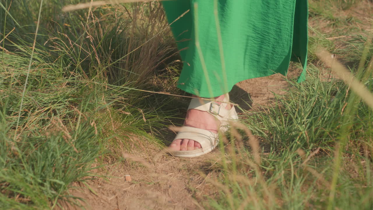 Close up of lady feet in sandals walking downhill through dry grassy hillside wearing green gown, with soft warm sunlight highlighting natural movement and texture of grass and footpath